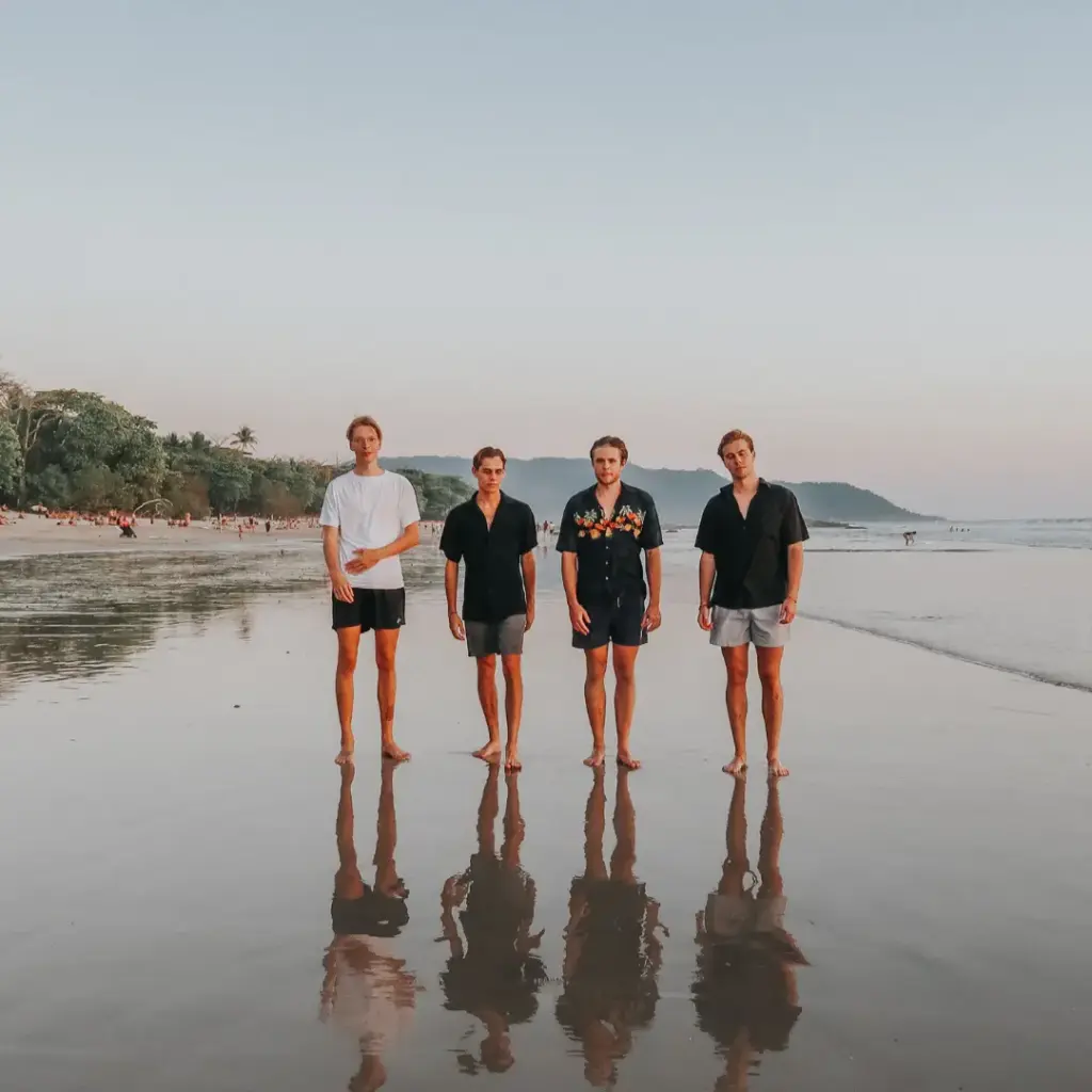Four young men standing barefoot on a wet beach at sunset, reflected in the sand, for why the name Gospel Saturated