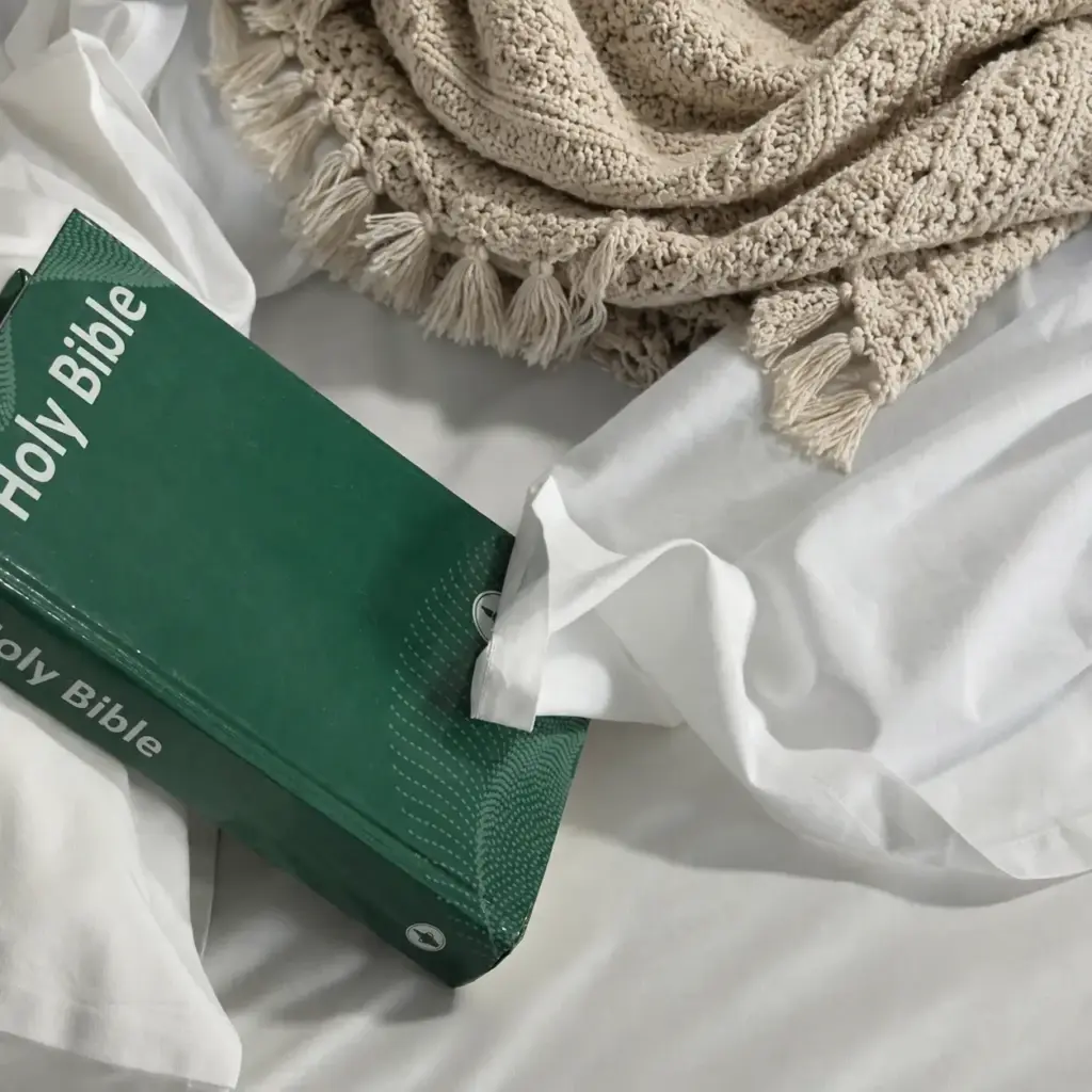 Green Holy Bible resting on white bed linen beside a cream knitted throw, illustrating how we write at Gospel Saturated